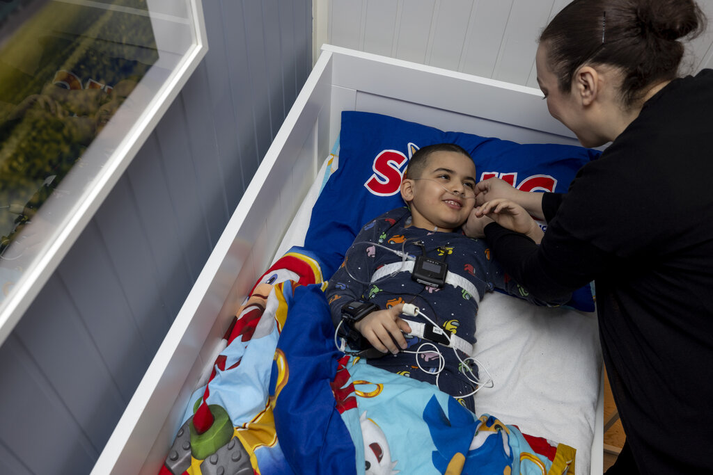 The photo shows a boy lying in his bed. Tools for sleep testing and monitoring are equipped on the boy. His mother is bending towards him, both are smiling
