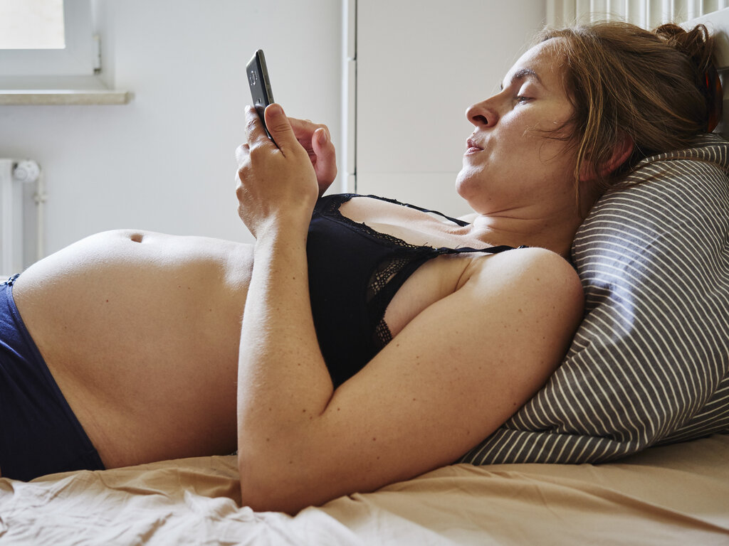 Bildet viser en gravid kvinne som ligger i BHen på senga. Hun kikker på mobilen og ser litt misfornøyd ut