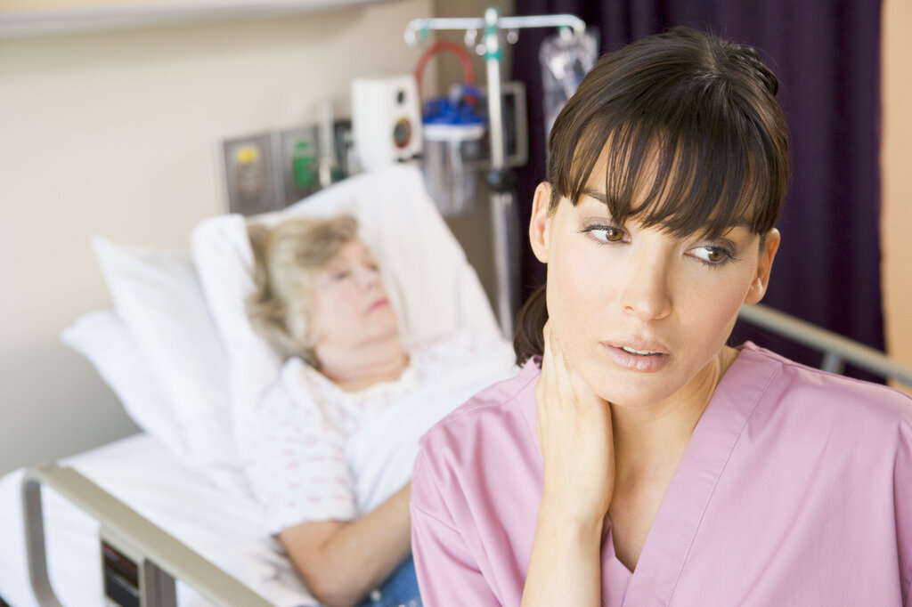 Bildet viser en sykepleier med en pasient i bakgrunnen som ser veldig sliten ut.