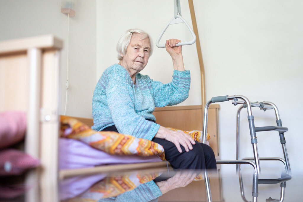 Gammel dame på pleiehjem sitter på sengen med rullator foran. Rutete pledd. 