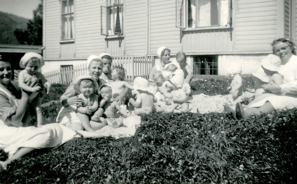 Historisk, gammelt bilde av sykepleiere med små barn på gresset utenfor Bergen kommunale mødrehjem.