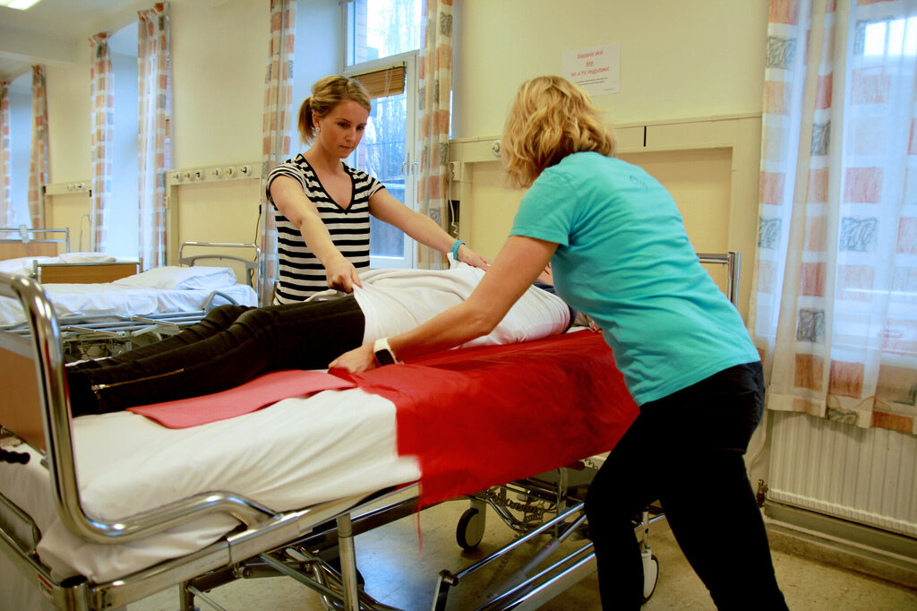 Bildet viser bedriftsfysioterapeutene Anne Aschehoug Loftu (t.v.) og Sigrunn Haug Simensen vise hvordan man kan bruke stikklaken og forflytningsduk ved forflytning av pasient i seng.