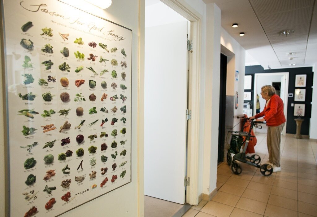 Bildet viser en gang på Rundskuedagens plejecenter med en plakat av grønnsaker på veggen, en kvinnelig beboer i bakgrunnen.