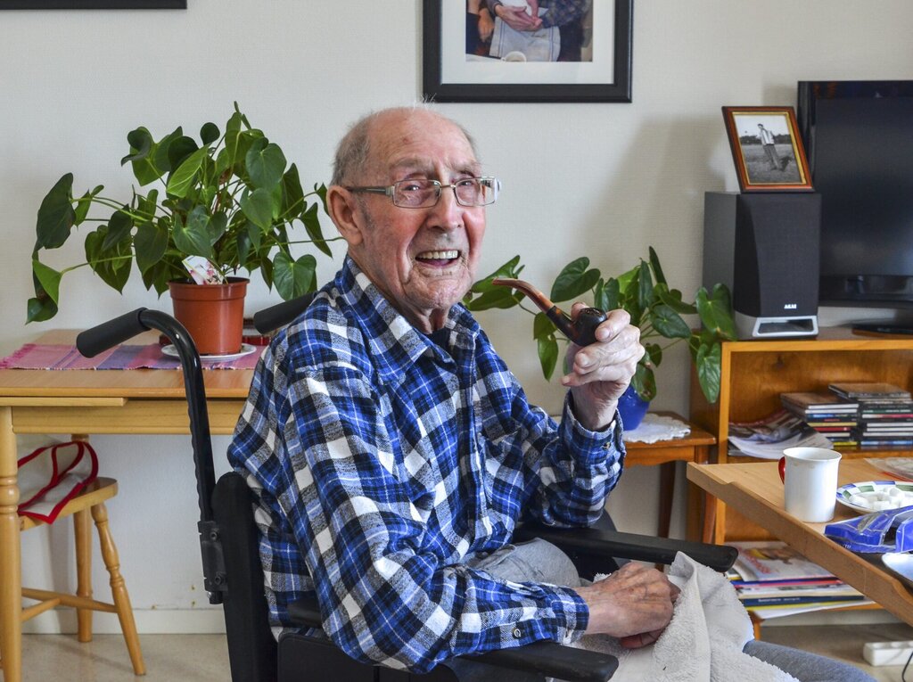 Bildet viser John Gundersen på 93 år. Han sitter i en rullestol i omsorgsboligen sin. Han har en pipe i hånden og smiler til fotografen.