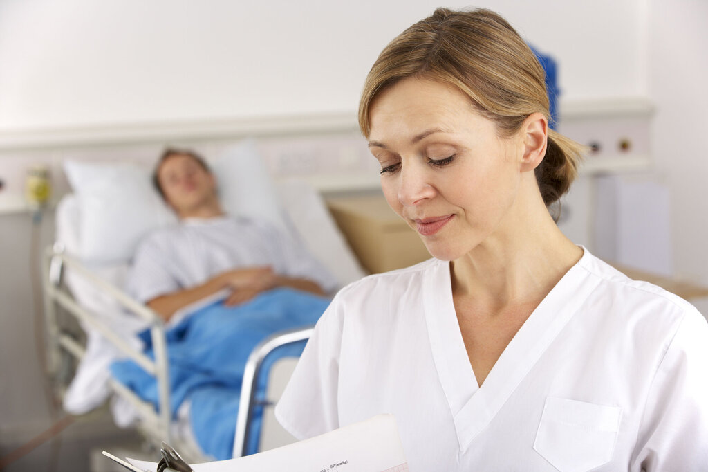 Bildet viser en sykepleier som står foran en seng på sykehuset med en pasient i.