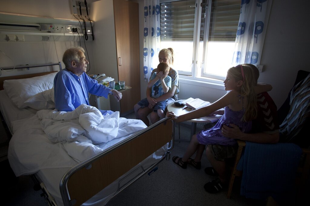 Bildet viser en syk, eldre mann i en sykehusseng. Han har besøk av barn og barnebarn.