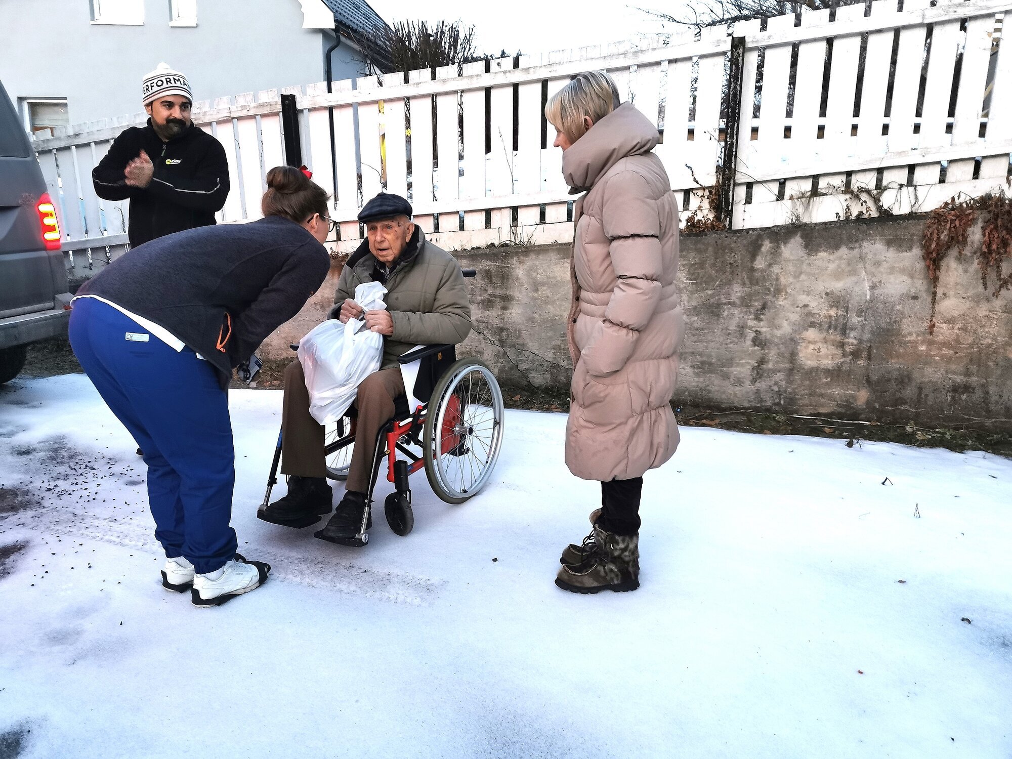 Mann i rullestol, sykepleier lener seg ned og prater. Pårørende til høyre, taxisjåfør til venstre. Vinter i hage.