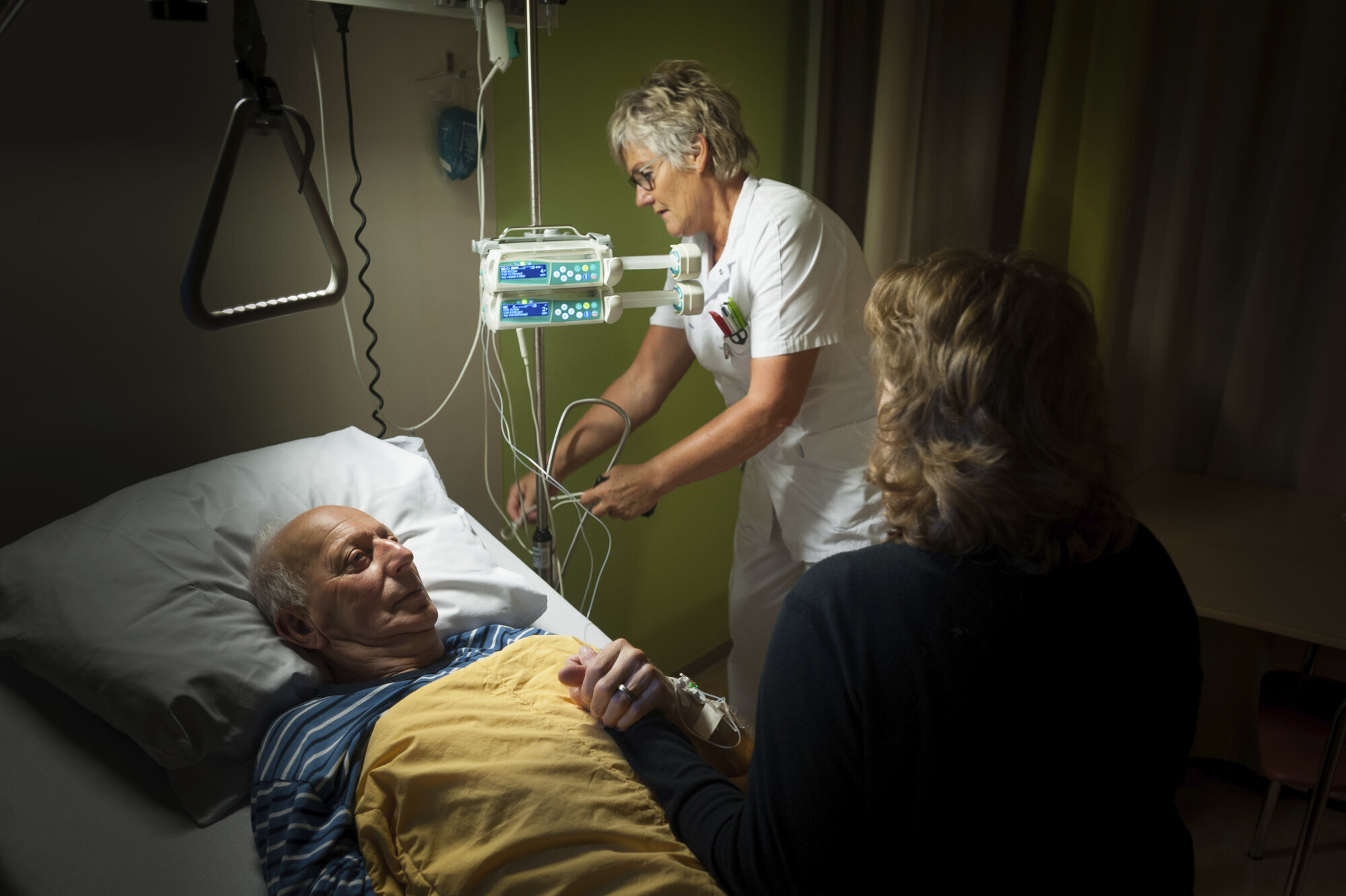 Eldre mann på palliativ avdeling Bildet viser en mann i sengen på palliativ avdeling. En sykepleier sjekker noen slanger, mens en pårørende kvinne sitter og holder mannen i hånda.