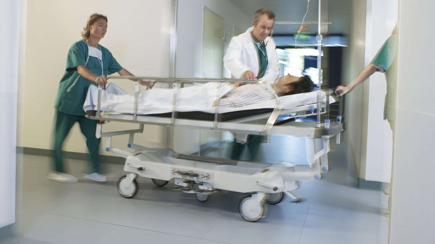 Pasient som trilles av helsepersonell Bildet viser en pasient i en sykehusseng som blir trillet av helsepersonell