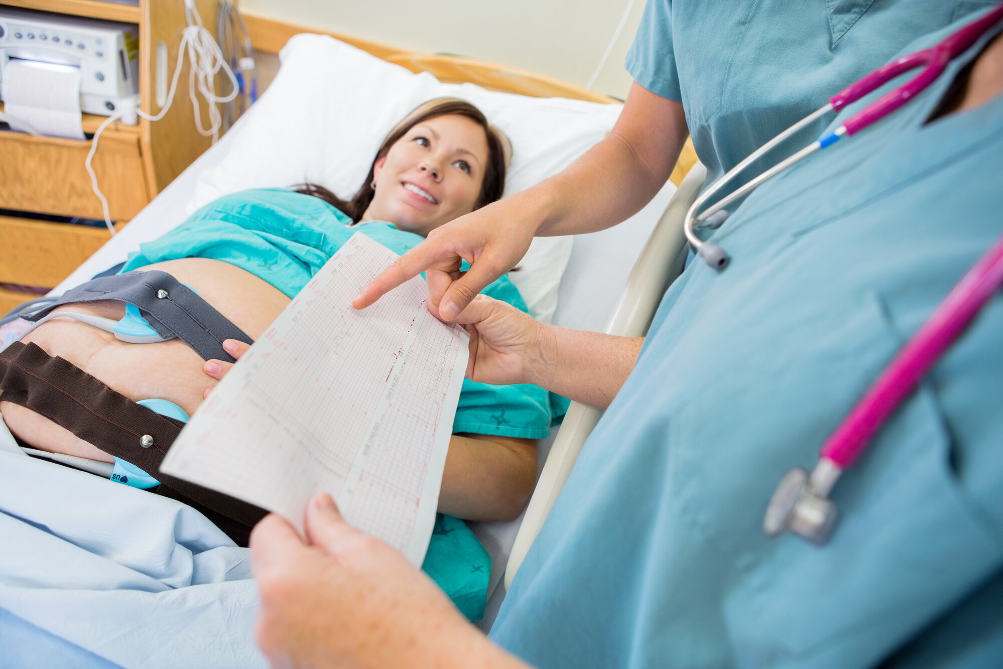 Høygravid kvinne og sykepleiere Bildet viser en høygravid kvinne som ligger i sengen og får ultralyd. To sykepleiere står ved siden av og diskuterer noe på et ark