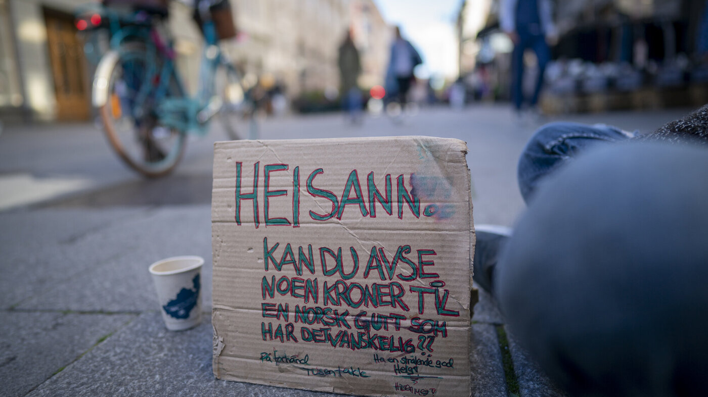 Bildet viser Karl Johans gate og en plakat som en sittende mann har foran seg, hvor det er bønn om bidrag til en som har det vanskelig.