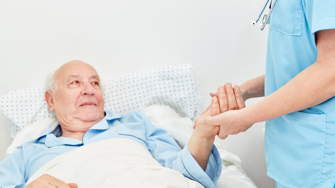 Bildet viser en eldre mann som ligger i senga. En sykepleier står ved siden av og holder ham i hånda