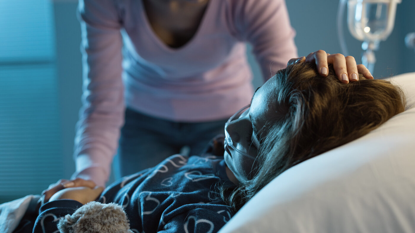 Bildet viser ei syk jente i senga på sykehuset. Moren står over og holder henne på pannen.