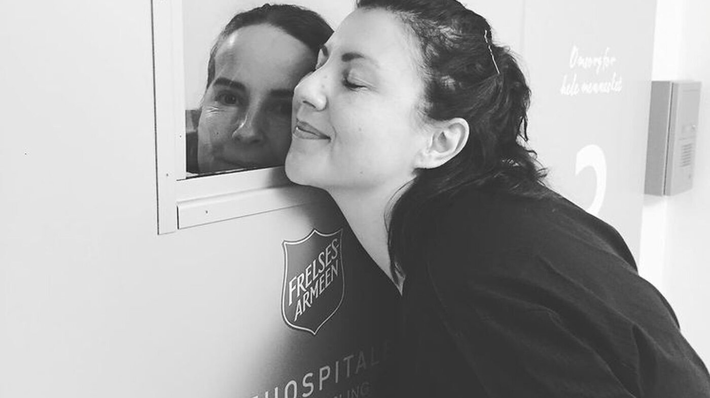 Bildet viser to sykepleiere som gir hverandre en klem gjennom en dør.