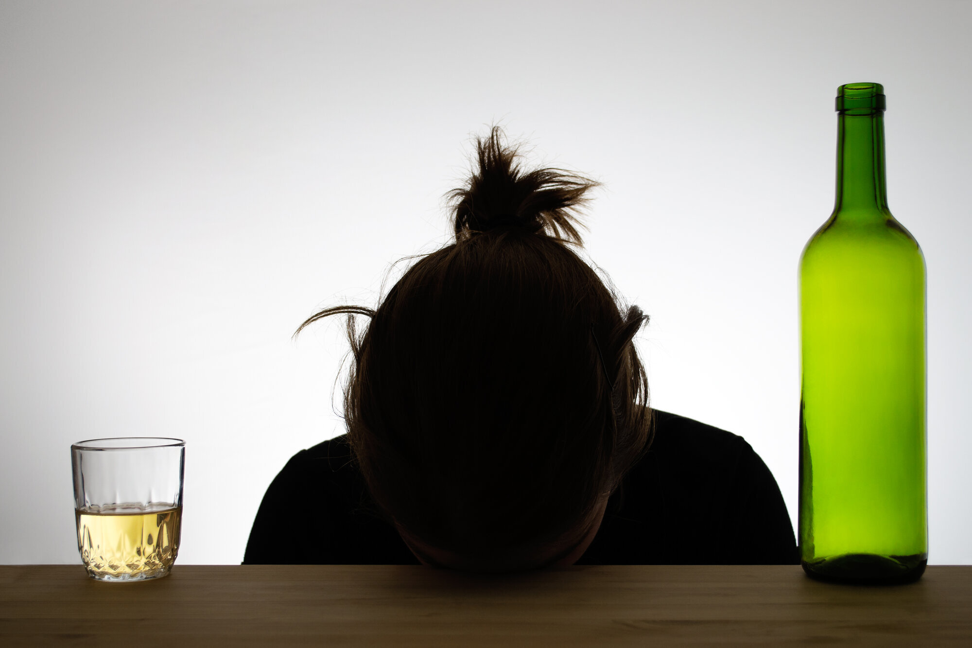 Alkohol og rus Bildet viser silhuetten av en kvinne som hviler hodet på en bordkant. Ved siden av henne står et glass med alkohol og en flaske.