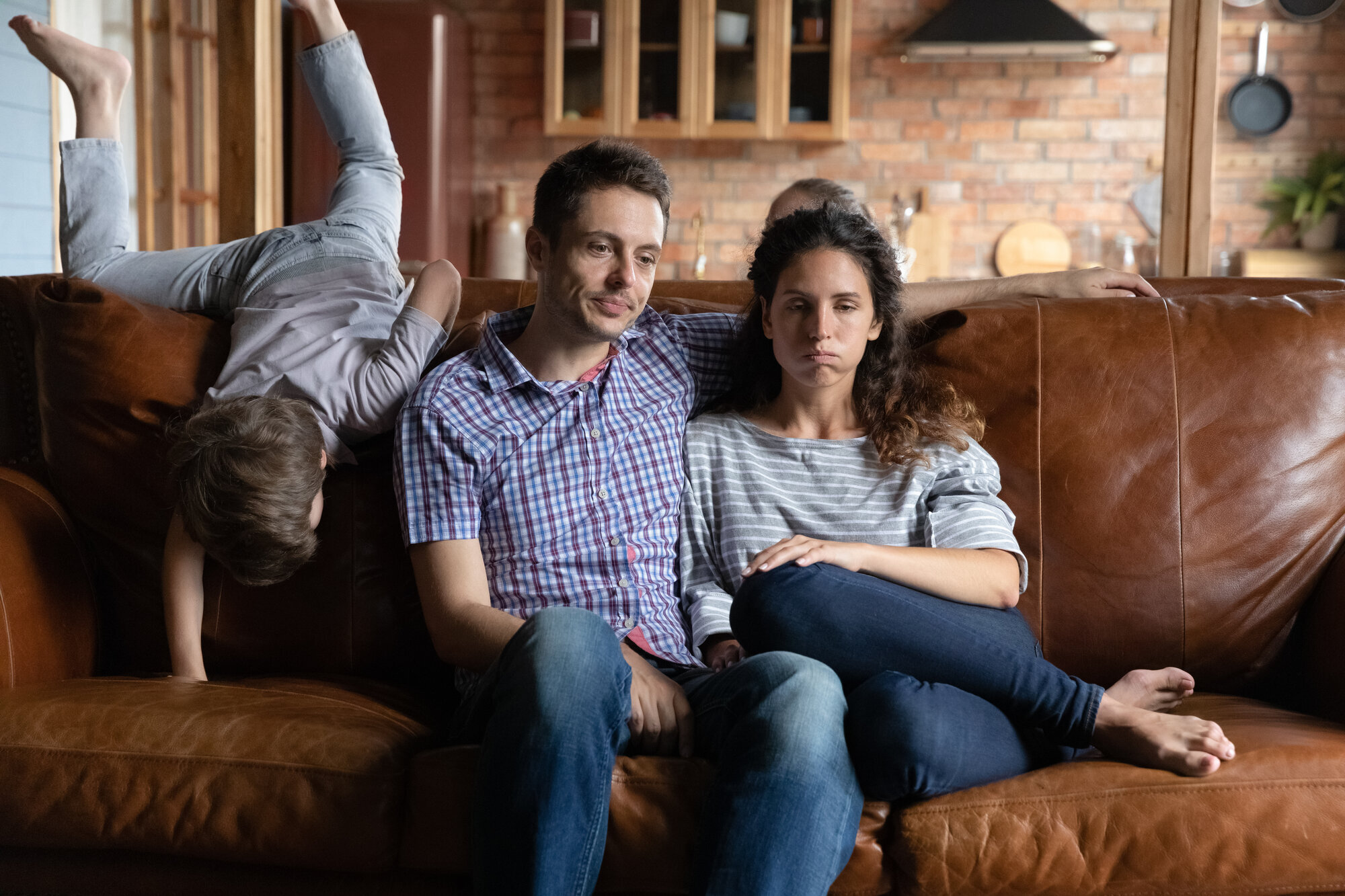 Familie Bildet viser en mann og en kvinne som ser slitne ut og sitter i en sofa, og et barn som klatrer på sofaryggen