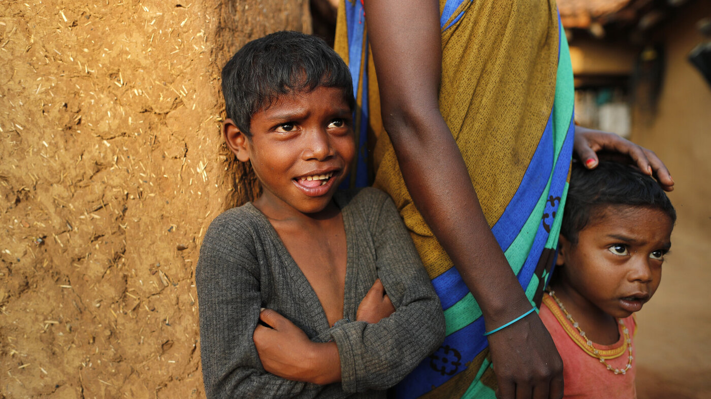 Bildet er fra India og viser to barn som står sammen med en voksen person og ser på helsearbeidere som overtaler eldre til å vaksinere seg mot korona