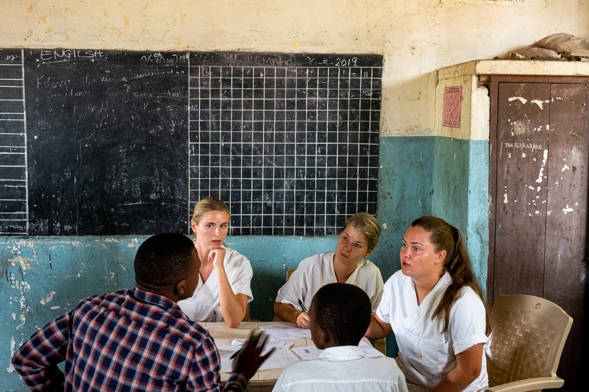 Sykepleierstudenter i Tanzania Bildet viser tre sykepleierstudenter fra Norge som er i Tanzania. De sitter og snakker med to afrikanske menn.