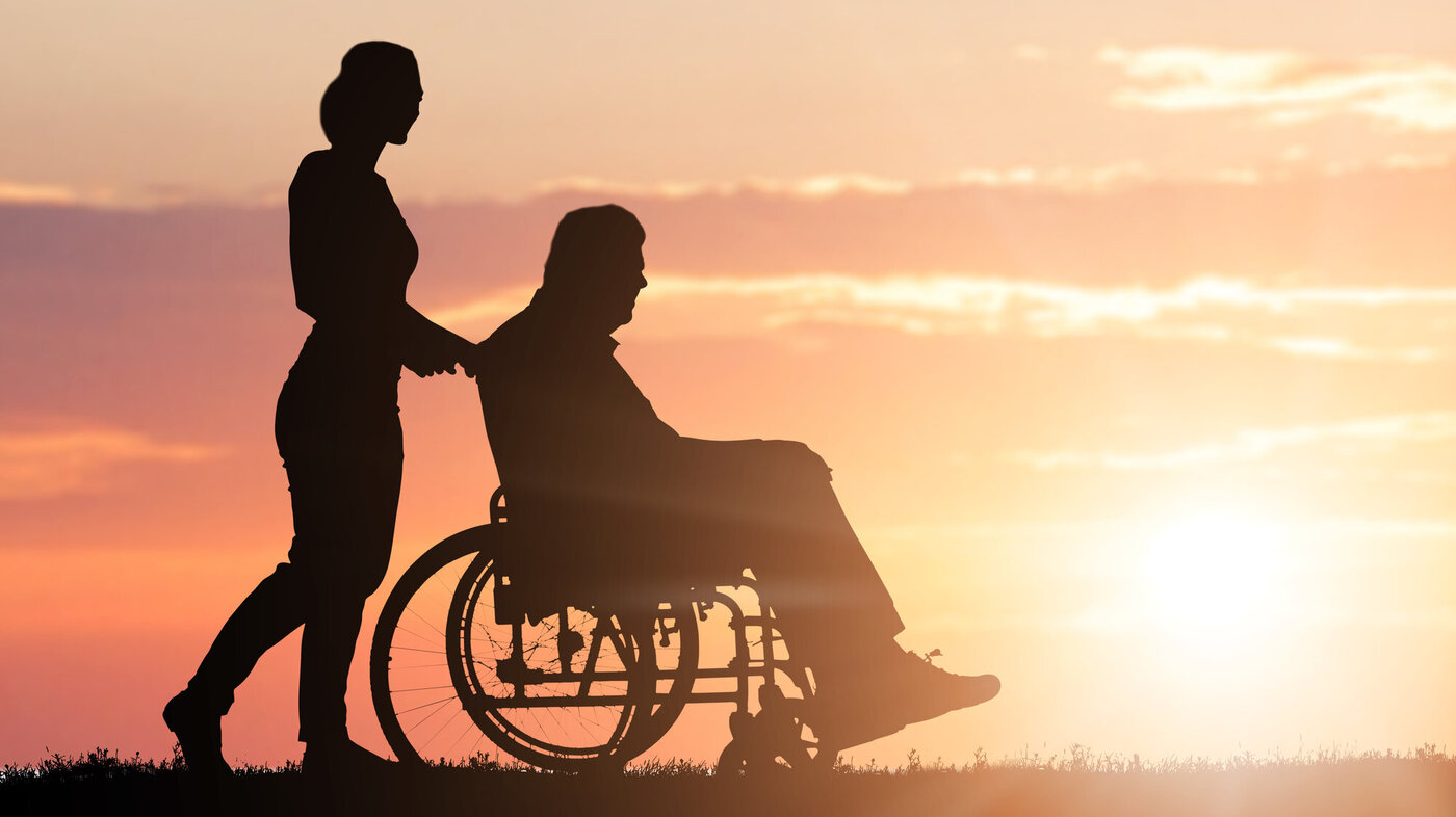 Bildet viser silhuetten av en kvinne som triller en eldre mann i rullestol i solnedgang.