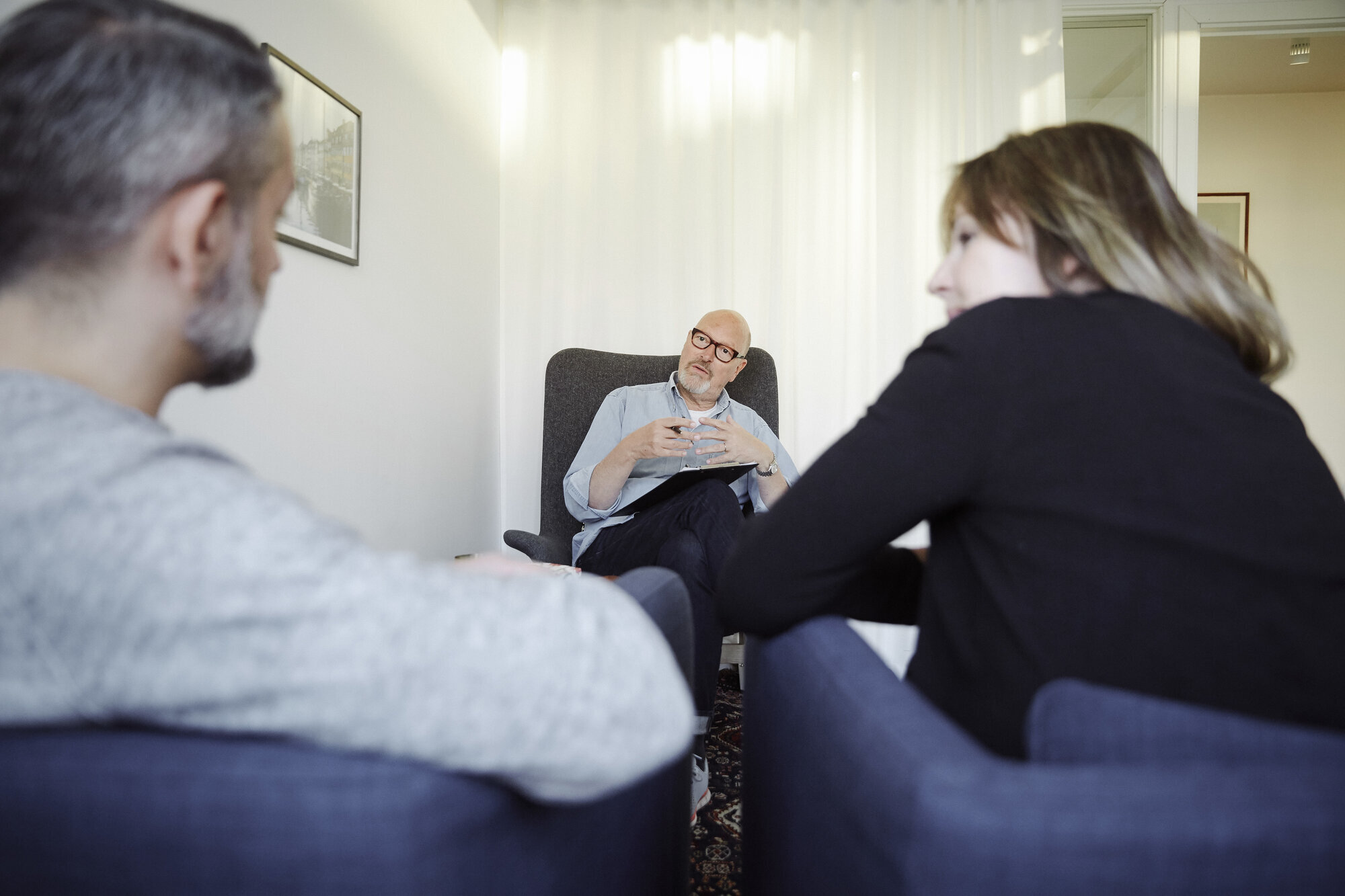 Terapisituasjon Bildet viser en mannlig terapeut som er vendt mot leseren. Foran ham sitter en mann og ei dame, som vi ser ryggen på.