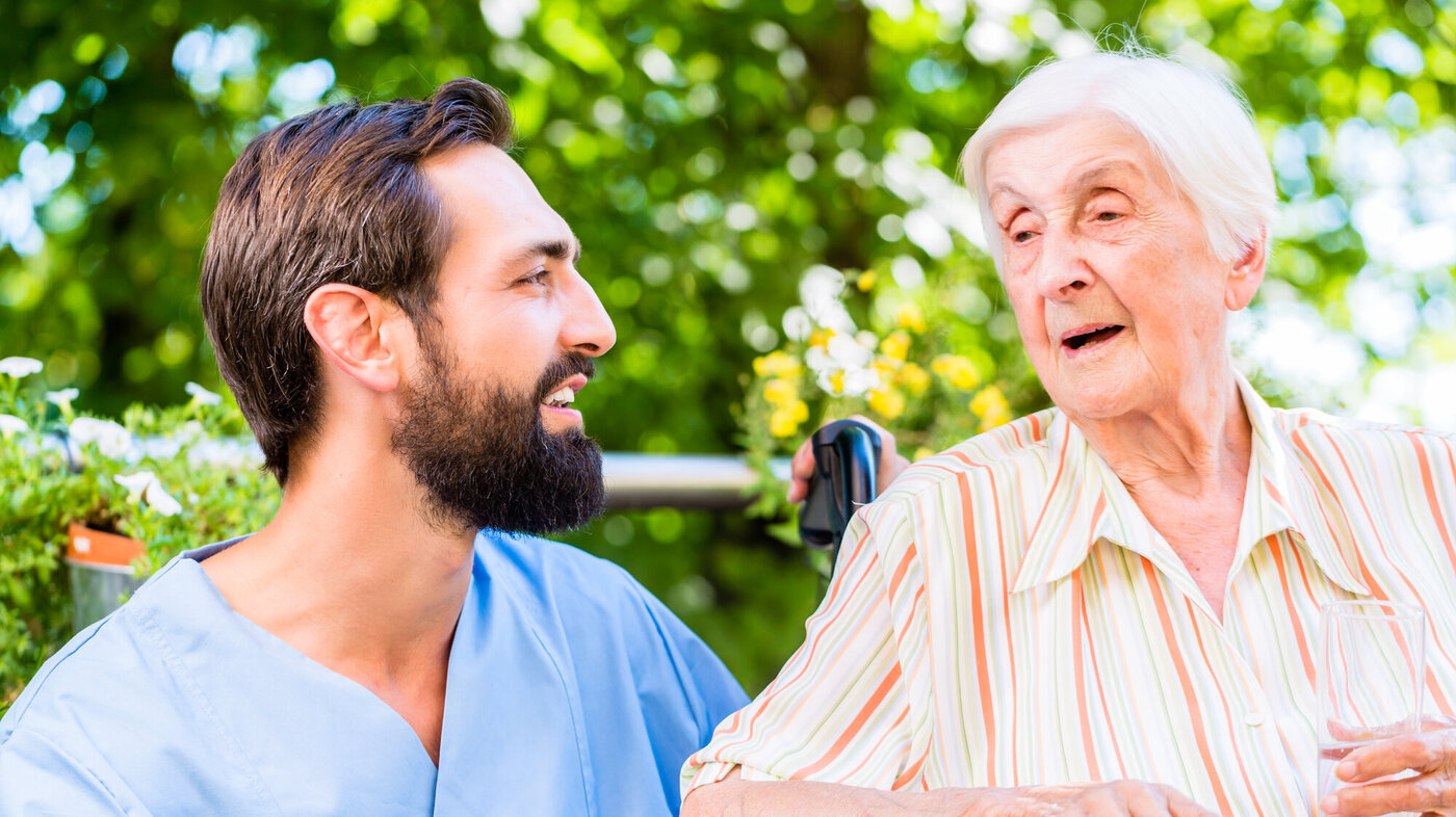Bildet viser en mannlig sykepleier som snakker med en eldre dame. De sitter ute, og det er god stemning.