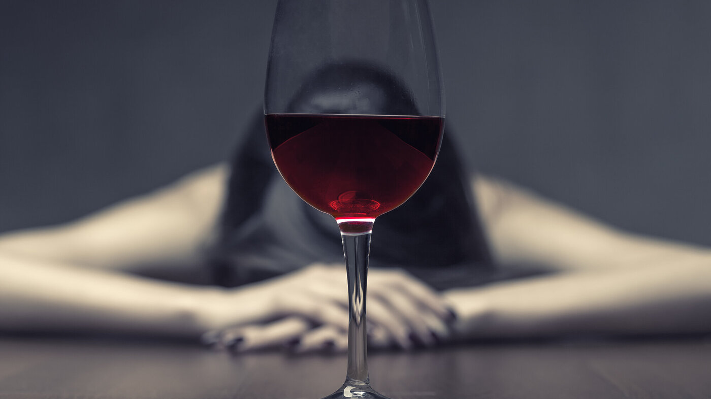 Bildet viser en kvinne som hviler hodet på et bord. Ovenfor henne står et glass rødvin