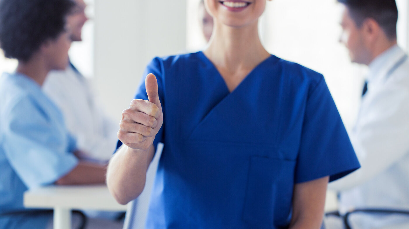 Bildet viser en sykepleier i front som viser "tommel opp" mens hun smiler.