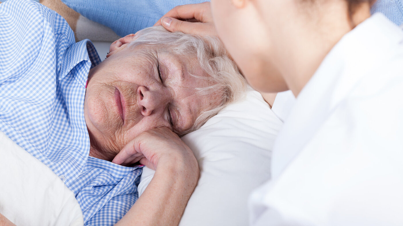 Bildet viser en syk dame i sengen. En yngre dame sitter ved sengekanten og stryker henne over håret