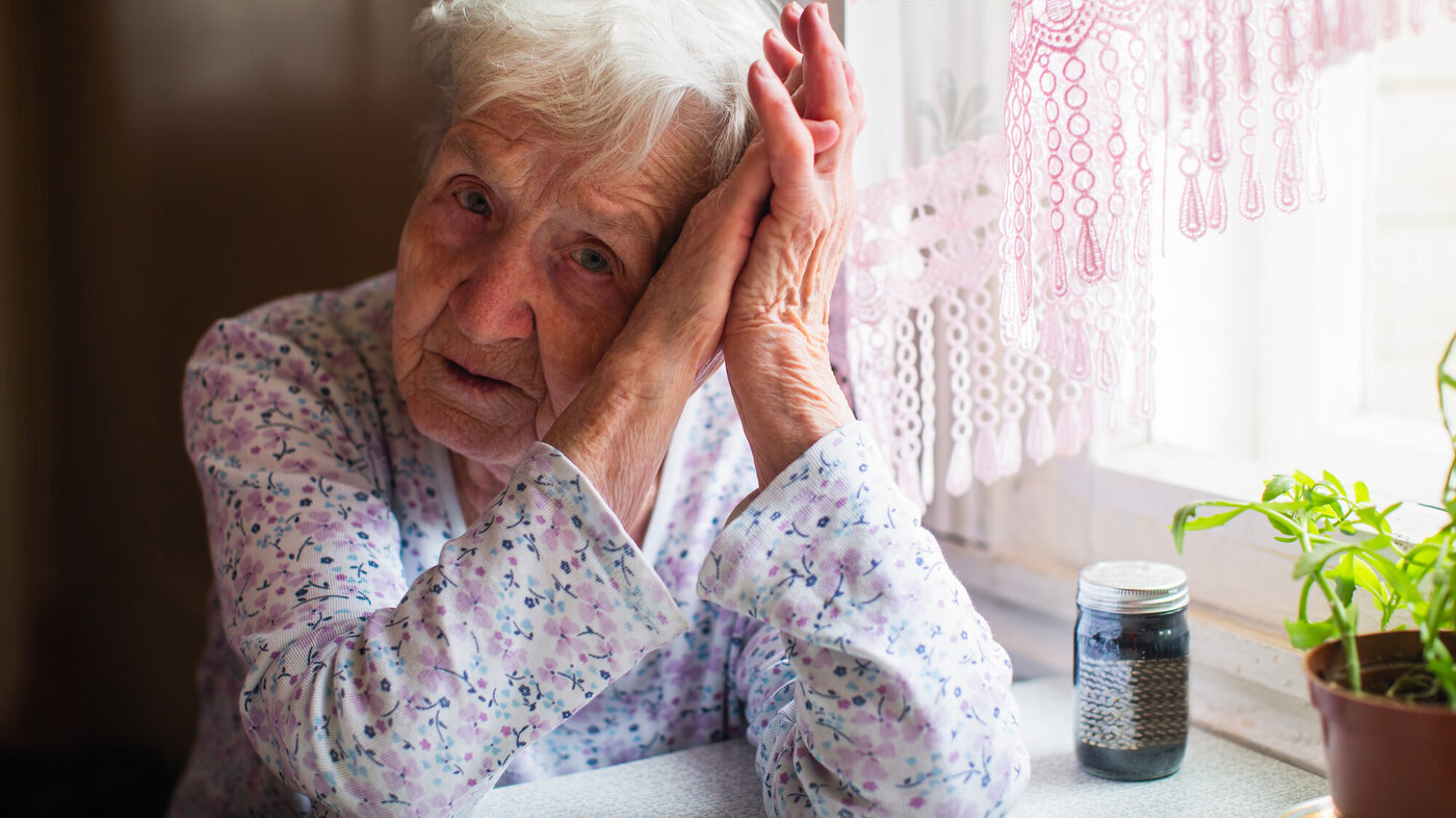 Skrøpelig eldre dame Bildet viser en trist, gammel dame som lener hodet mot hendene sine. Hun sitter ved kjøkkenbordet med et pilleglass foran seg.