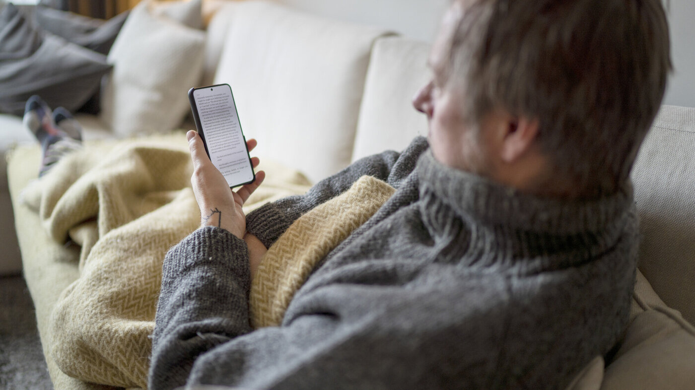 Bildet viser en mann som ligger under et pledd på sofaen og ser på mobilen