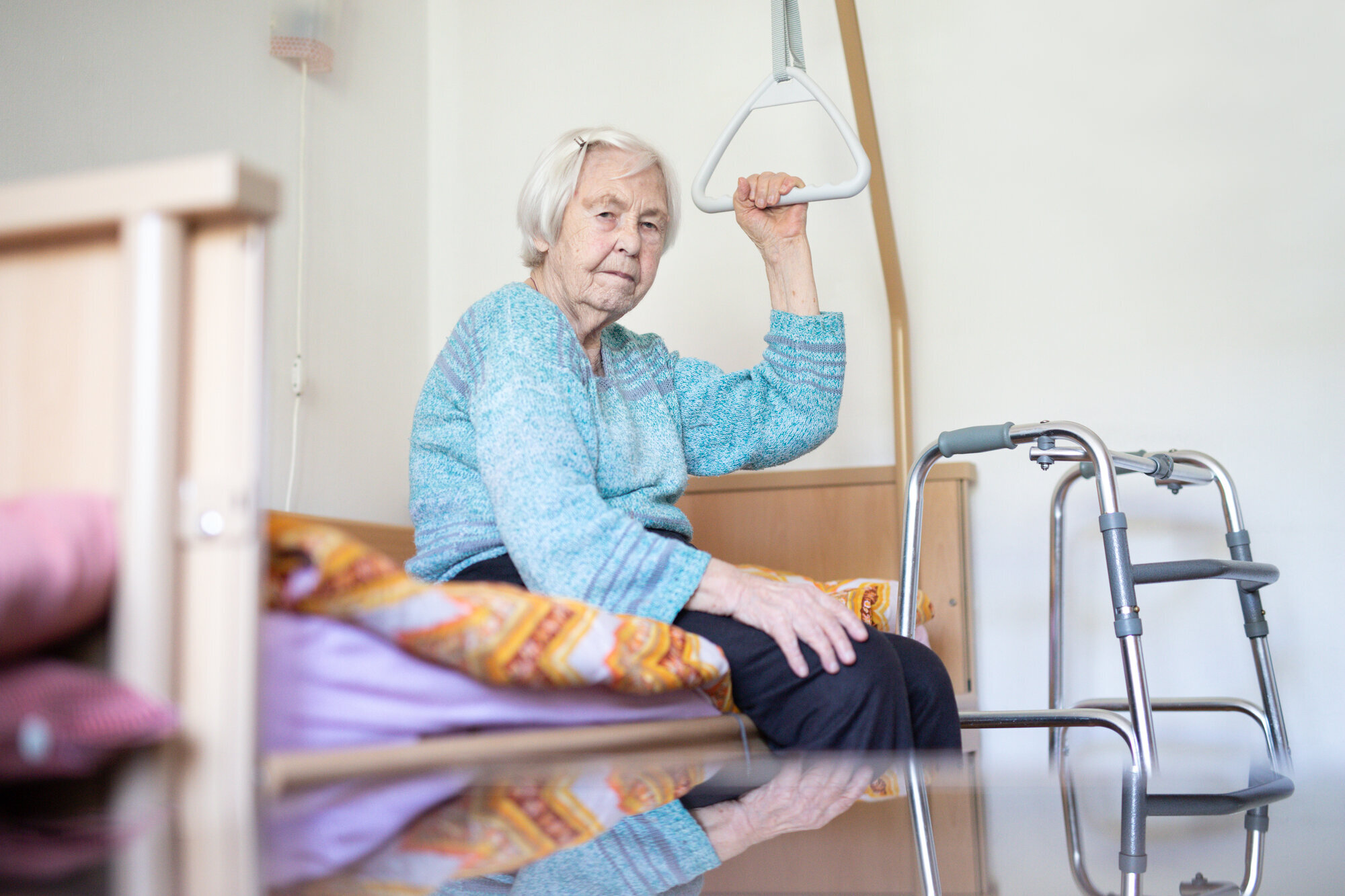 Gammel dame på pleiehjem sitter på sengen med rullator foran. Rutete pledd.