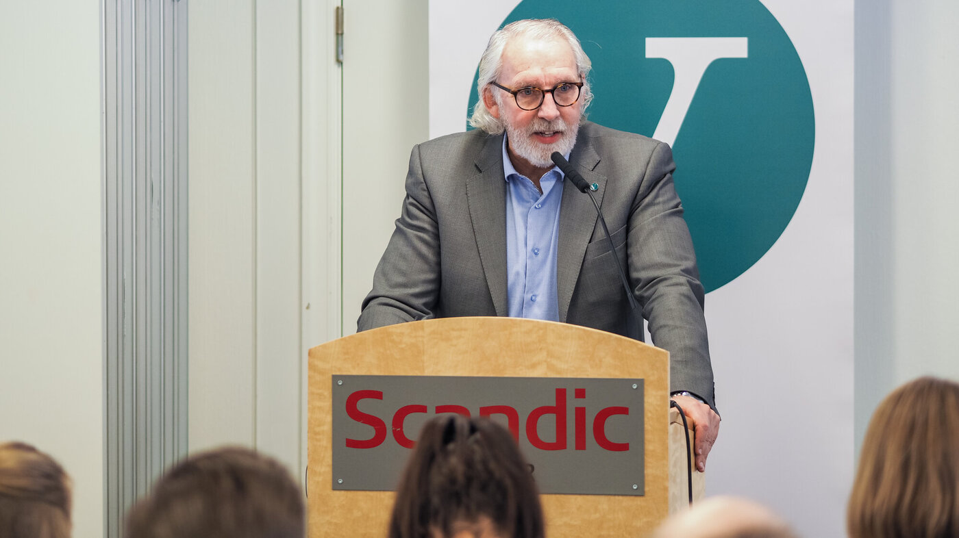 Senior mannlig politiker på talerstol i stort lokale med Venstres logo i bakgrunnen