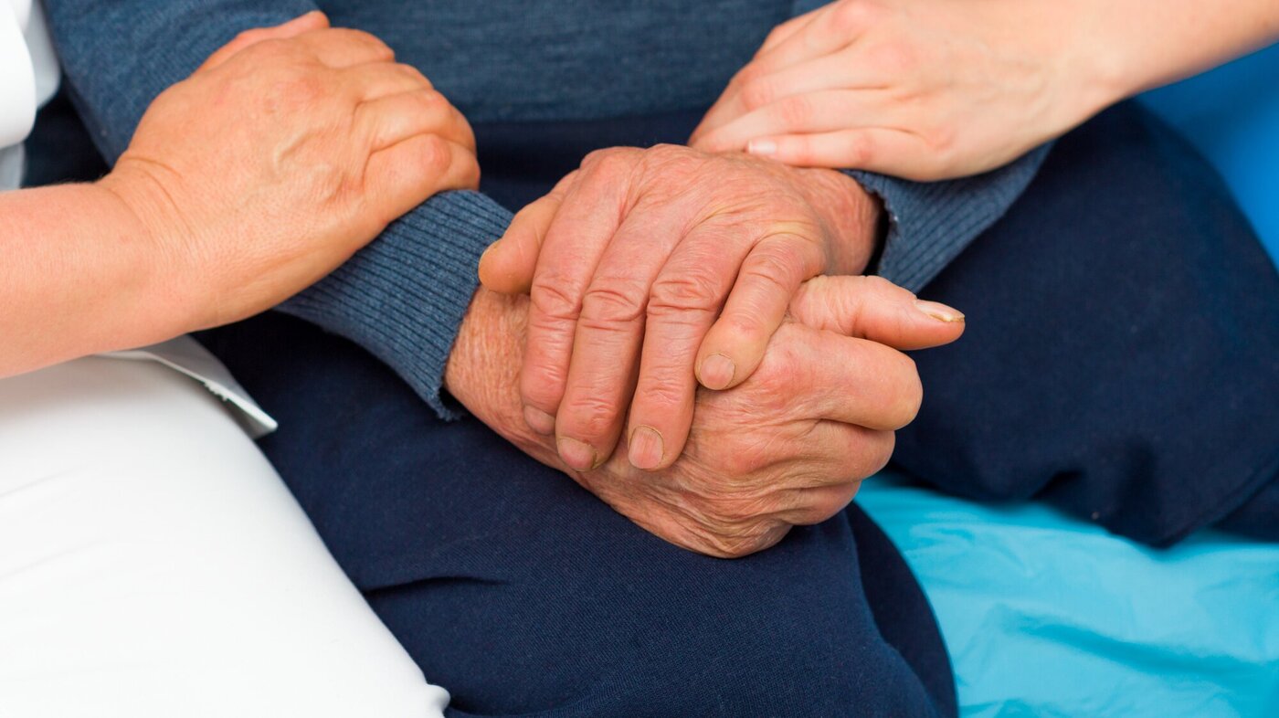 Pasientens to hender er foldet i fanget, mens to hender til pleiere holder varsomt på armene til pasienten. 