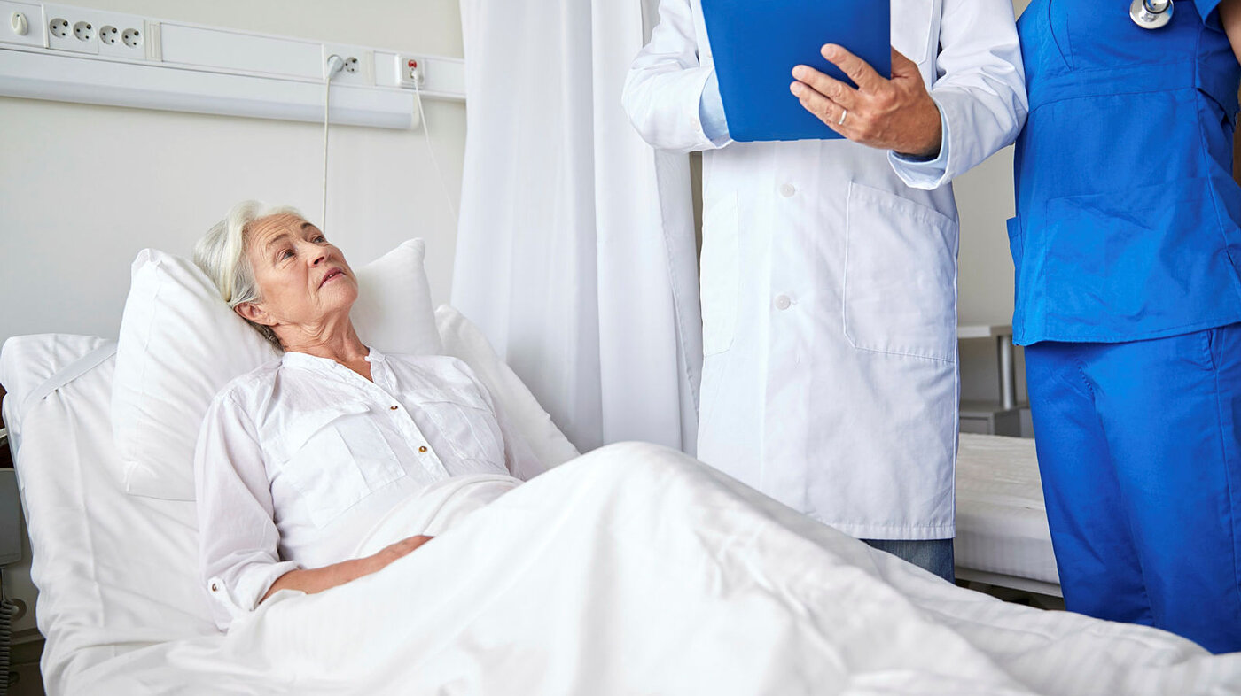 Bildet viser en syk, eldre dame i en sykeseng. Ved sengen står en lege og sykepleier på visitt.