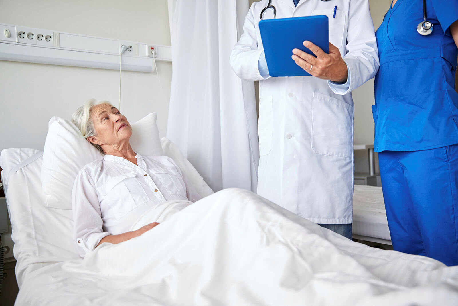 Lege og sykepleier på visitt hos syk dame Bildet viser en syk, eldre dame i en sykeseng. Ved sengen står en lege og sykepleier på visitt.