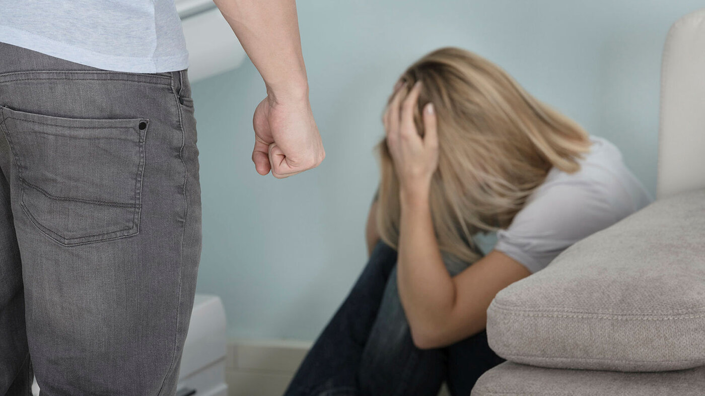 Partnervold Bildet viser en mann som står med knyttneven foran en redd dame som har krøket seg sammen bak sofaen.