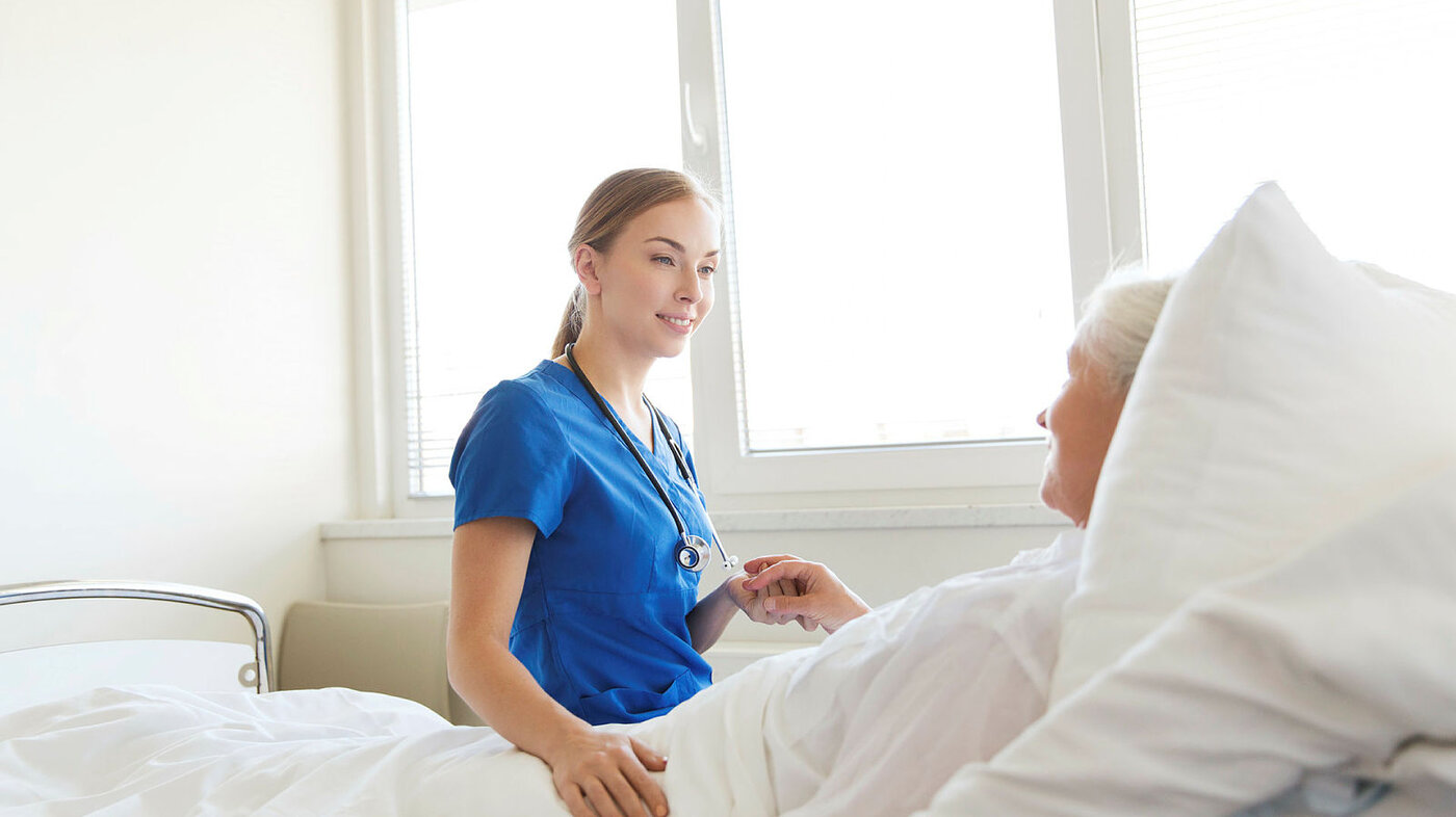 Sykepleier sitter på sengekanten til eldre, sengeliggende kvinnelig pasient.