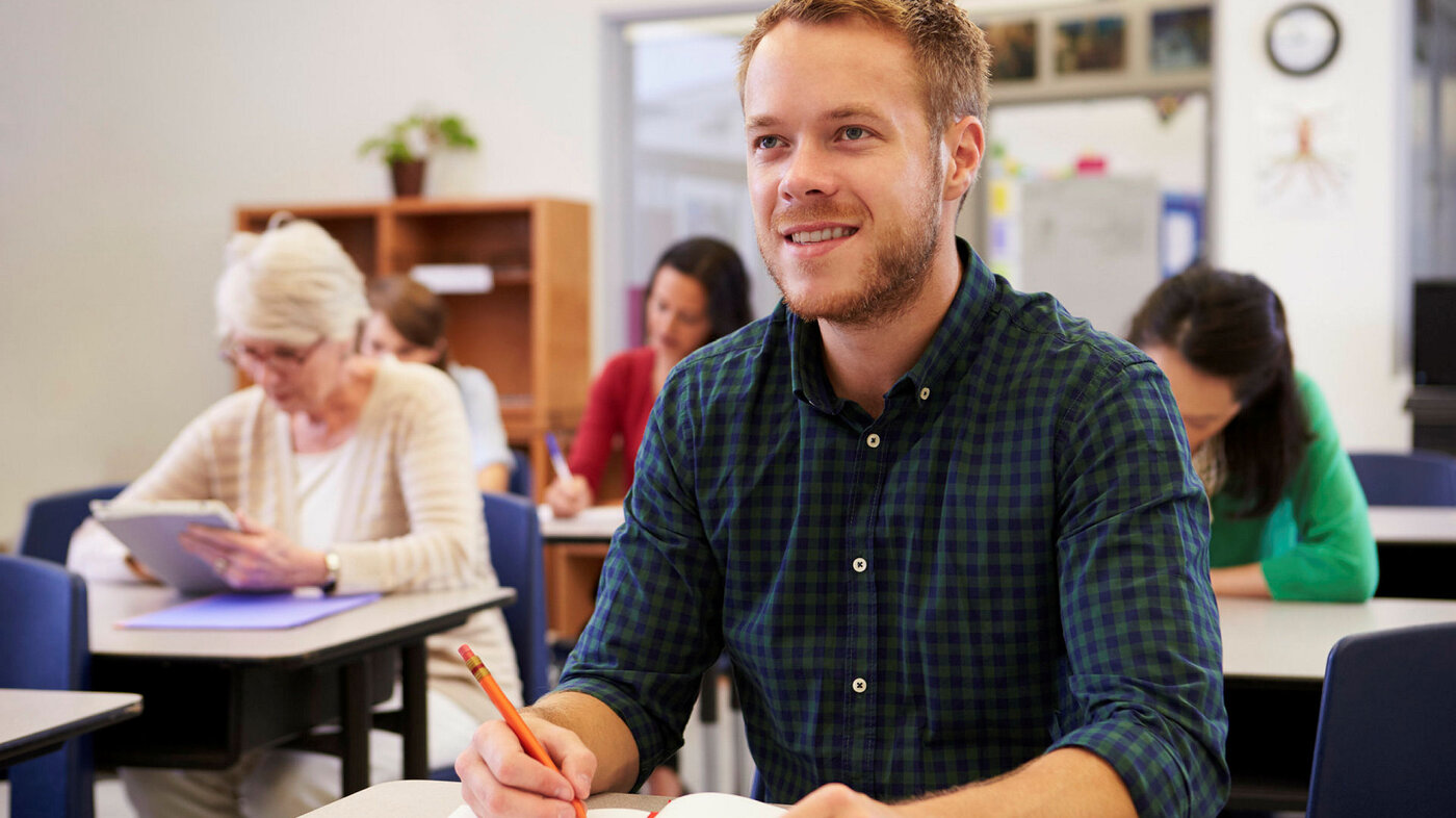 Klasseromsundervisning Bildet viser en ung mann i et klasserom. Det er flere studenter også i bakgrunnen.