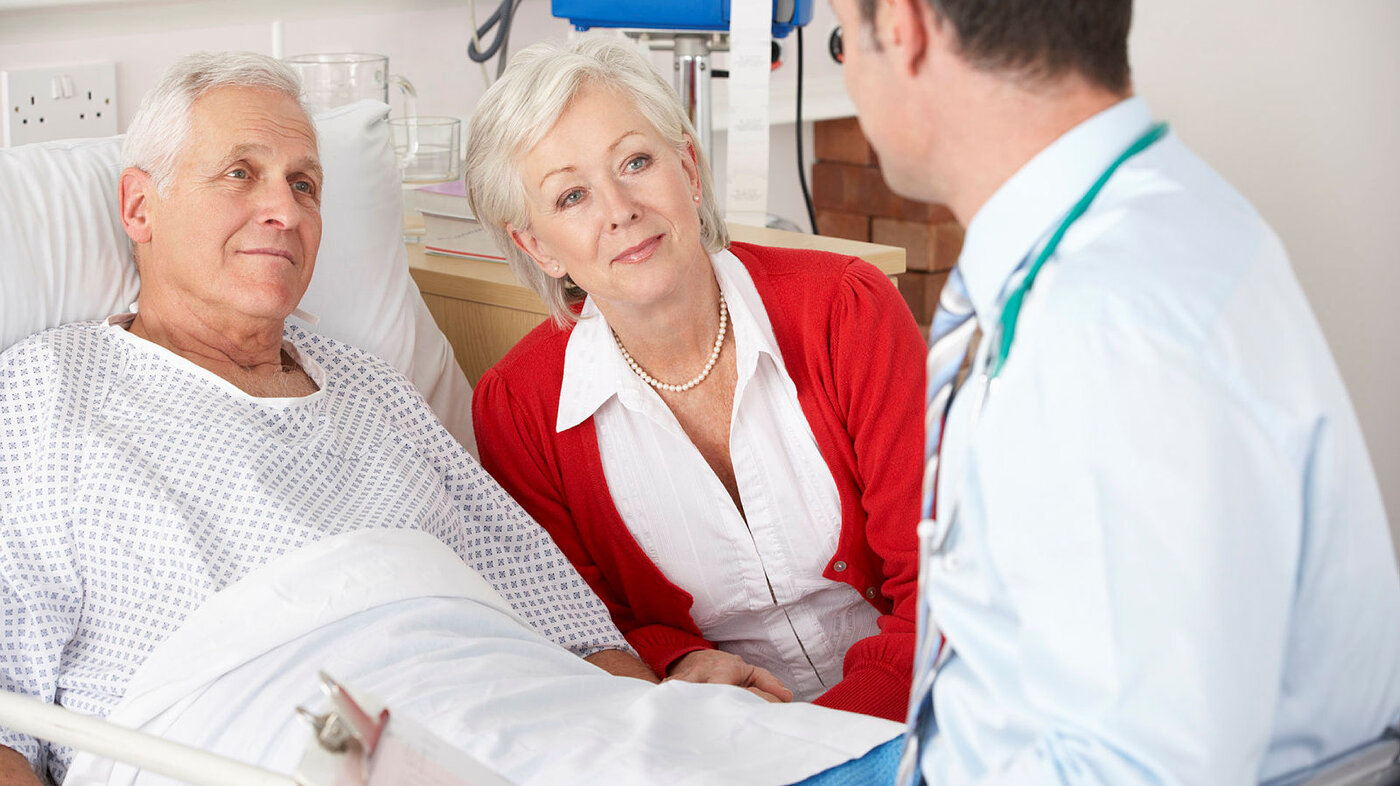 Bildet viser en eldre mann som ligger i senga på sykehuset. Kona sitter ved siden av, og legen er på visitt.