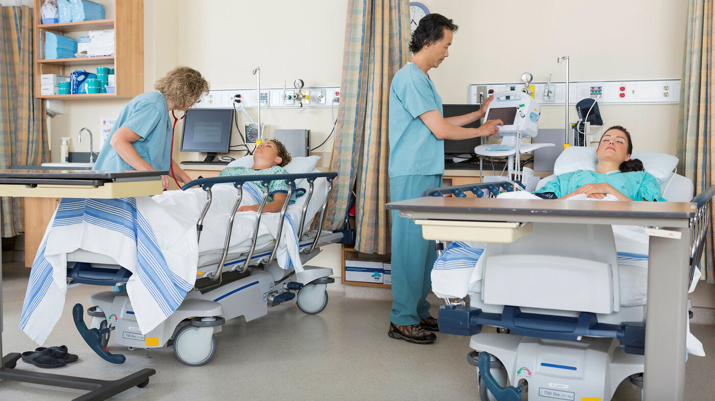 Bildet viser to pasienter og to sykepleiere som står ved sengene deres på postoperativen.
