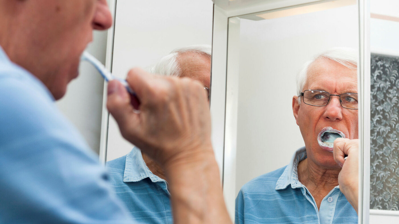 Bildet viser en eldre mann foran et speil som pusser tennene.