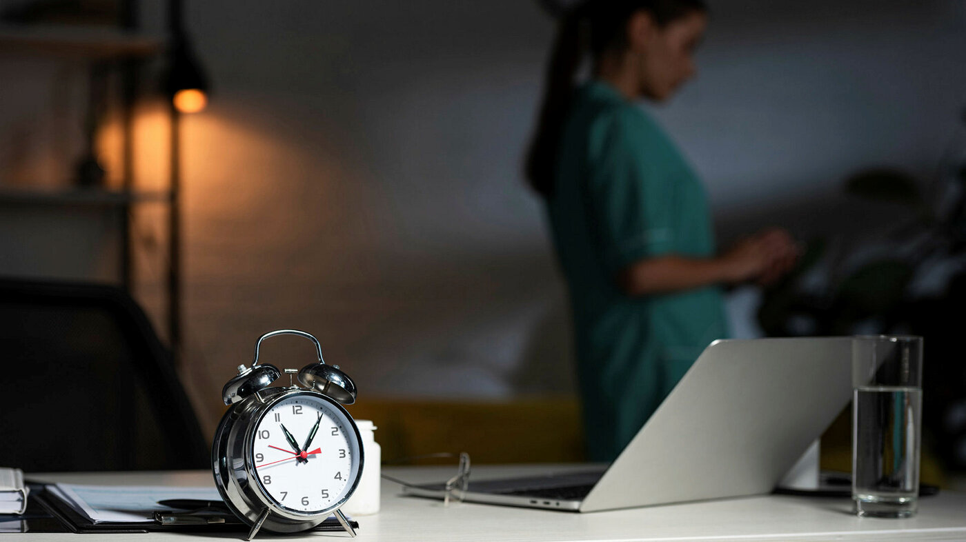Bildet viser en vekkerklokke i forgrunnen og en sykepleier og en tent lampe i bakgrunnen. Rommet er ganske mørkt.