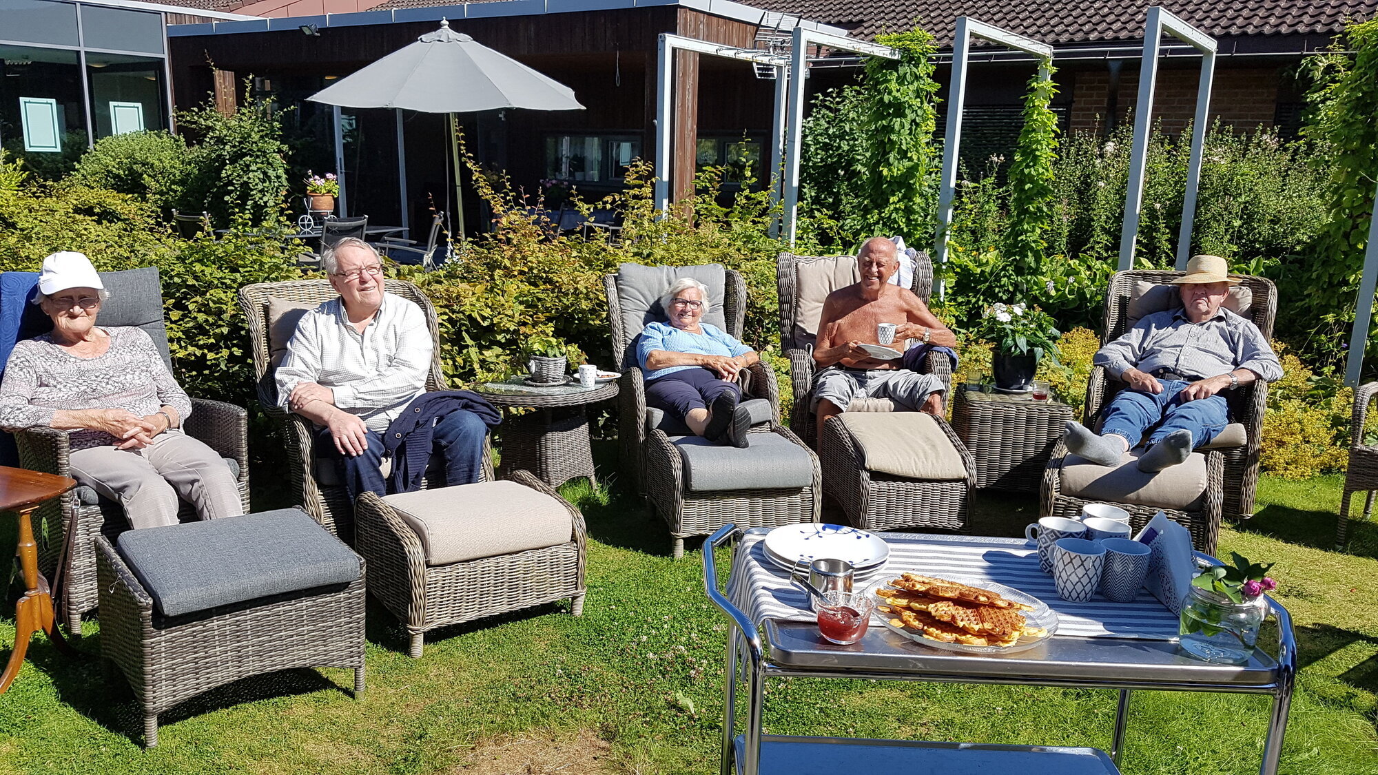 Sommerdag ved Haugsåsen bokollektiv i Skien Bildet viser sykehjemsbeboere som sitter i solstoler i hagen og nyter solen. Foran står et bord med vafler.