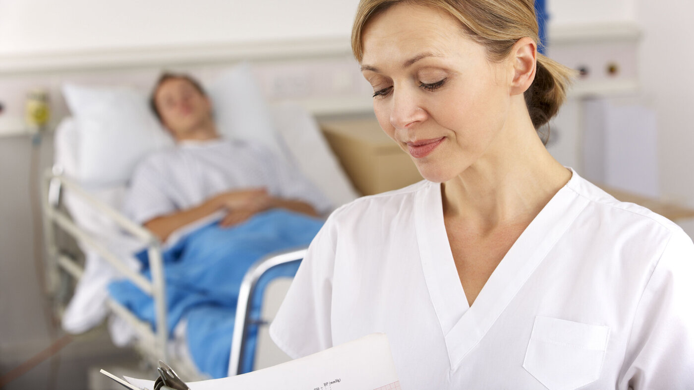 Bildet viser en sykepleier som står foran en seng på sykehuset med en pasient i.