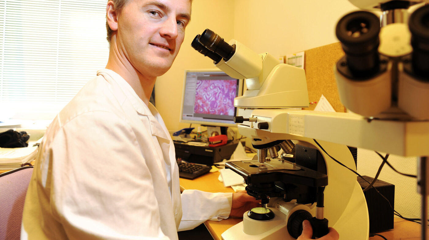 Bildet viser Sveinung Wergeland Sørbye som sitter ved et mikroskop, men ser inn i kamera.