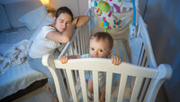 Bildet viser en mor som halvsover over barnesenga, mens barnet står i senga, lys våkent