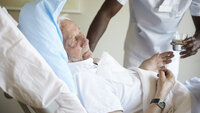Bildet viser en eldre mann som får vann av en mannlig sykepleier.