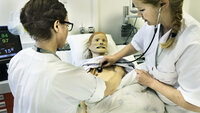 Bildet viser to sykepleiere som står ved en seng. I senga ligger en simuleringsdukke. Den ene sykepleieren lytter etter hjerteslag på dukken