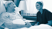Bildet viser en mannlig eldre pasient som ligger i en sykehusseng. En kvinnelig sykepleier sitter ved siden av sengen. Begge smiler