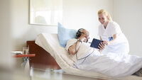 Bildet viser en kvinnelig sykepleierstudent som assisterer en eldre mannlig pasient som ligger i senga, med å bruke et nettbrett